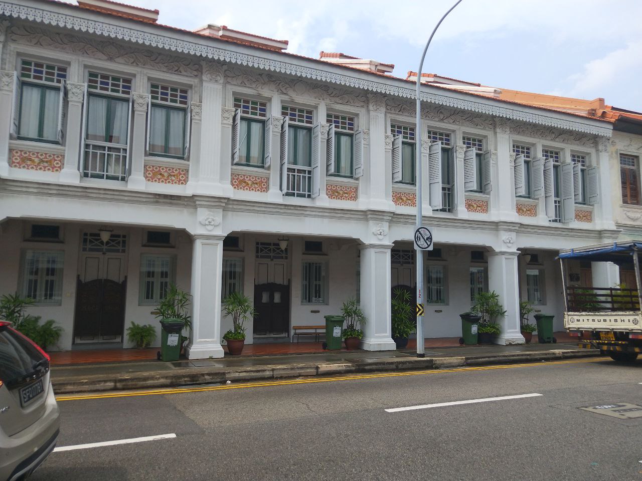 a street view in singapore showing an old white shophouse on the left side of the street. it is a twostory building with large windows on the second floor and doors on the first floor. there are green plants in pots in front of the building. a truck is parked on the right side of the road. the road has a yellow line painted on it. there is a sign on the post in front of the shophouse. there is a streetlight with a pole on the left side of the road.