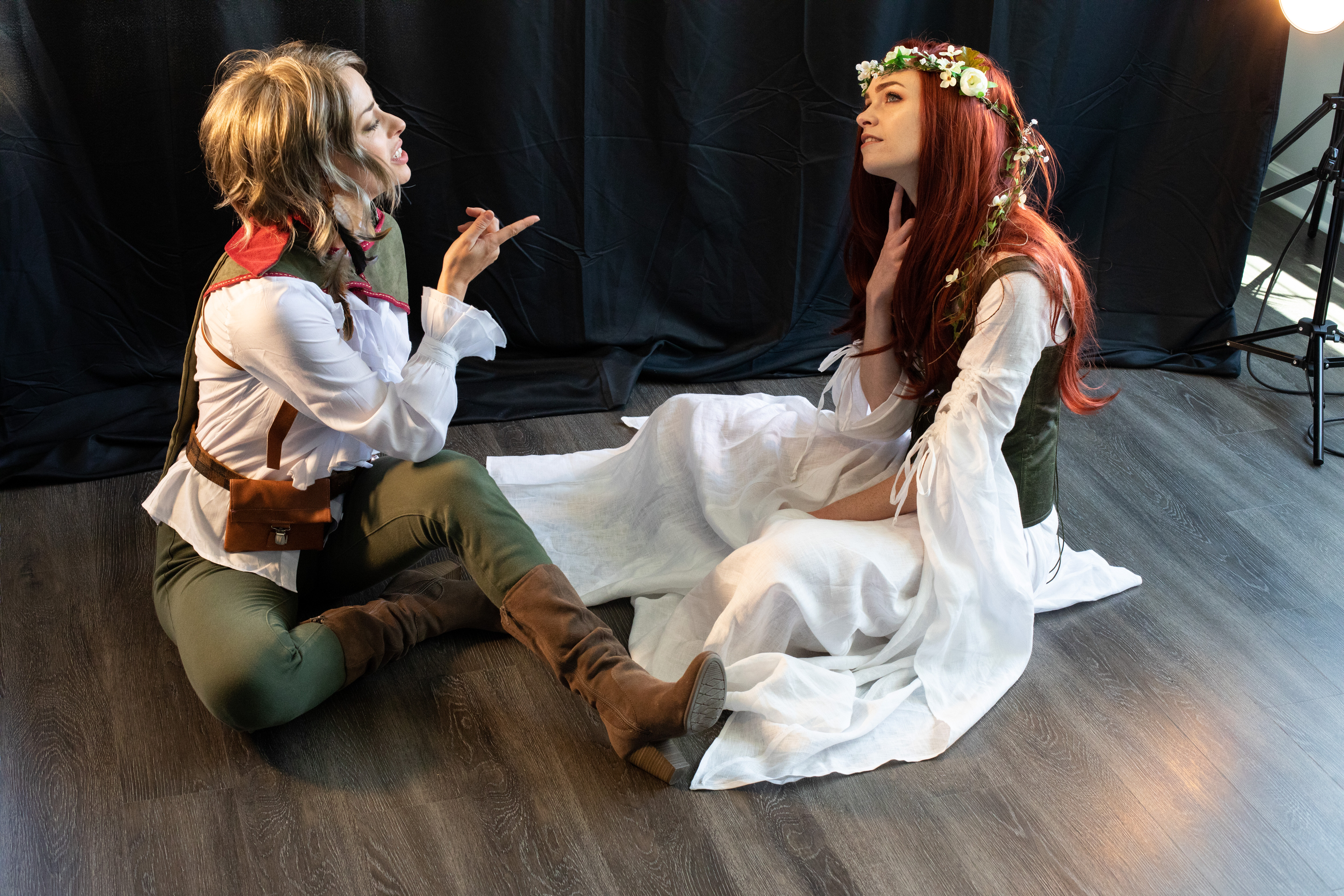 two women are cosplaying fantasy characters in an indoor setting. one woman, dressed in green and white, sits with her legs crossed while gesturing towards the other woman. the other woman, dressed in white and green with a flower crown, sits with her legs tucked underneath her and her head tilted back. the image appears to be taken from a photoshoot, as suggested by the backdrop and lighting.