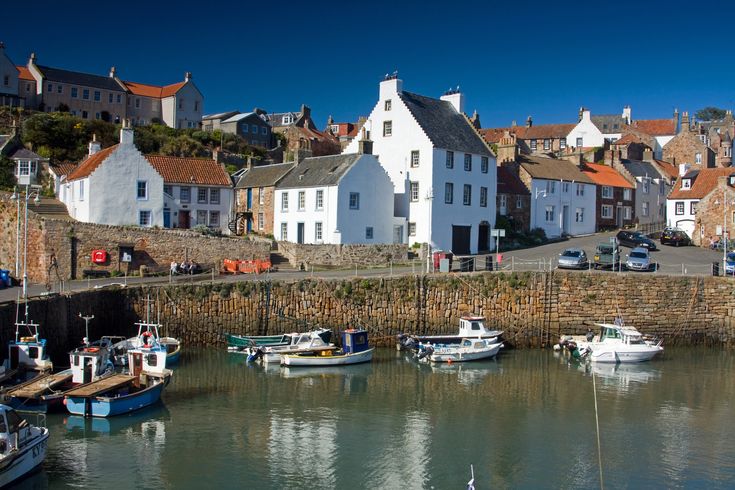 the image shows a picturesque view of the harbor in pittenweem, a charming coastal village in fife, scotland.  several small fishing boats are moored in the harbor, their hulls reflecting the clear, calm water. the harbor is enclosed by a sturdy stone wall. behind the wall, a cluster of traditional stone houses with whitewashed walls and red tile roofs climb a hillside, creating a charming and tightlypacked village setting. the sky is a clear, bright blue, indicating a sunny day. the overall scene is idyllic and evokes a sense of peaceful coastal life.