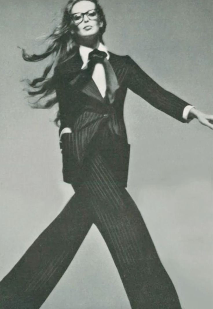 a black and white photo of a woman in a suit, with a tie and glasses, posing in a studio. her hair is long and flowing, and her pose is confident and stylish. the photo is a classic example of fashion photography from the 1970s.