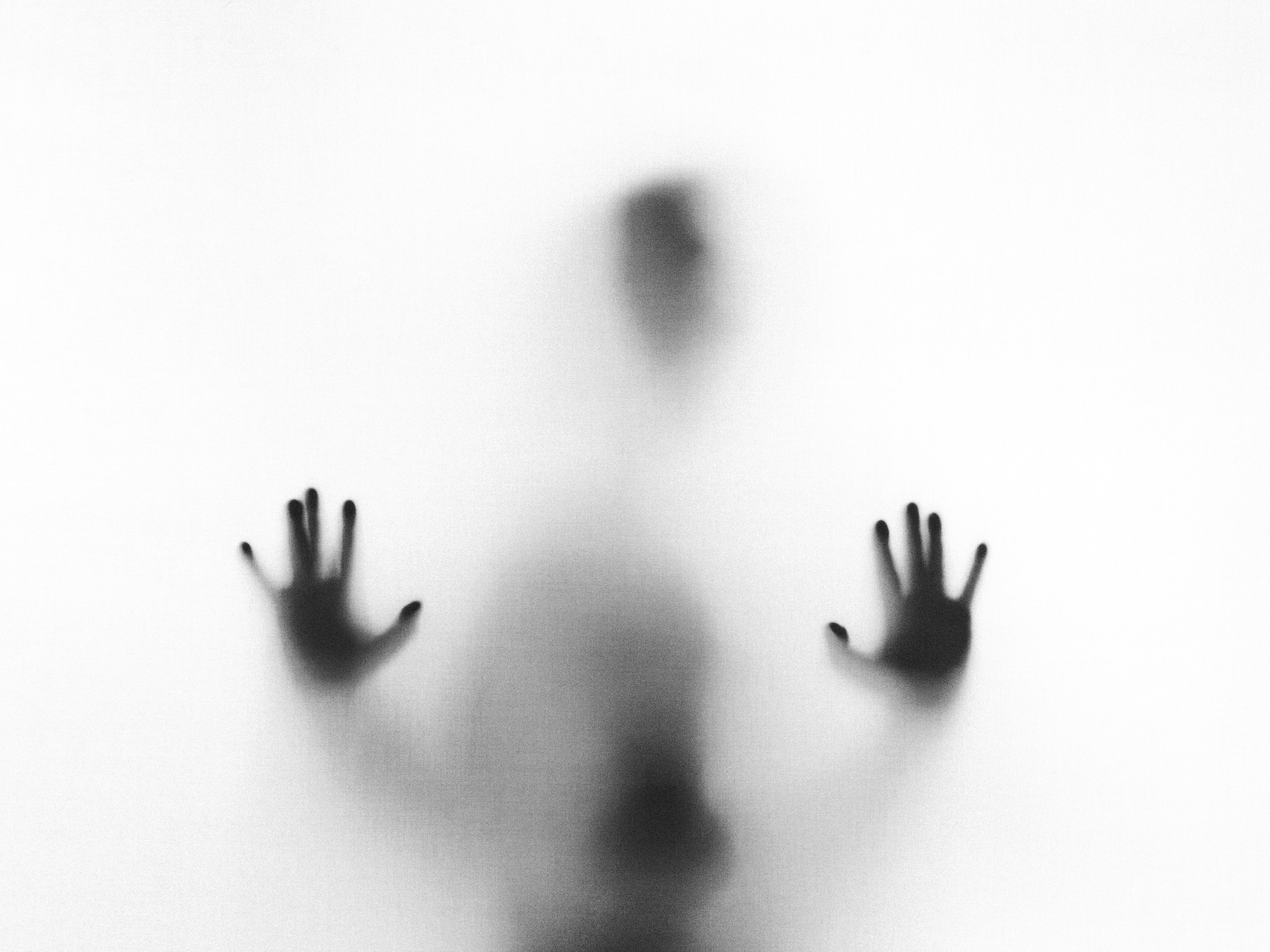 a shadowy silhouette of a person is seen through a translucent white surface, their hands pressed against it as if reaching out.  the figure is only vaguely defined, creating a sense of mystery and possible unease.