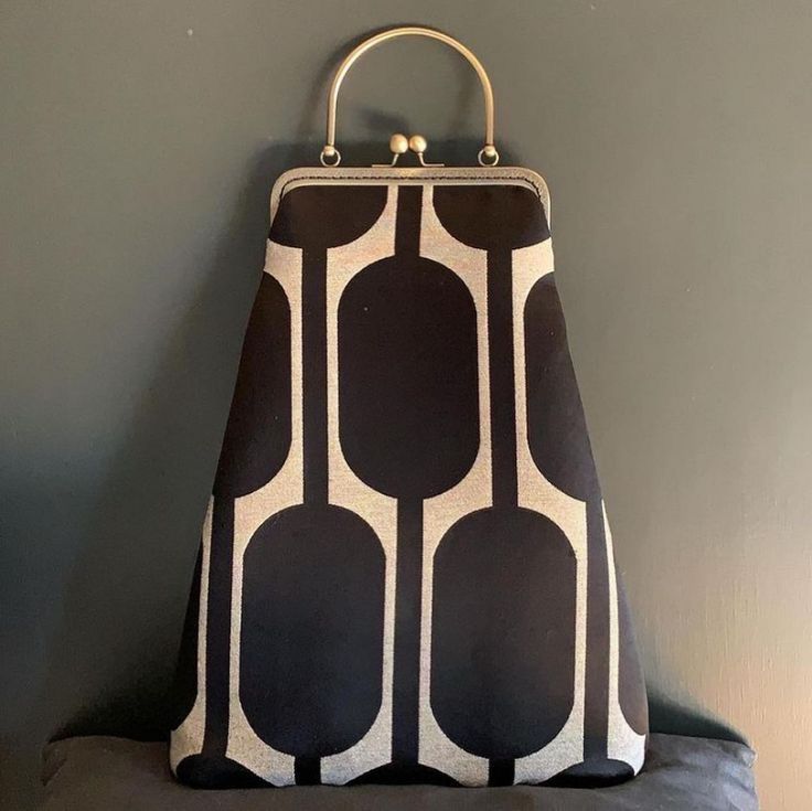 a black handbag with a retro geometric pattern of rounded rectangles stands against a dark background. the bag has a metal frame with a round handle and a clasp closure. the fabric has a black base with a lighter color pattern. it rests on a dark, soft surface.