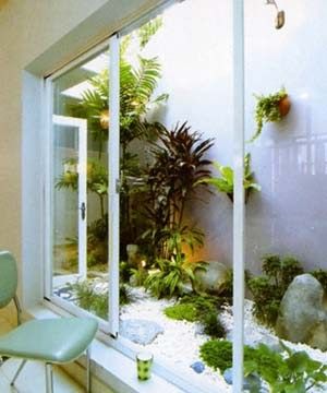 the image shows an indoor garden viewed through a large window. various plants and greenery fill the space, with large rocks and white pebbles scattered on the ground. a chair sits near the window, and a plant decorates the wall. the overall effect is a serene and natural environment brought indoors.