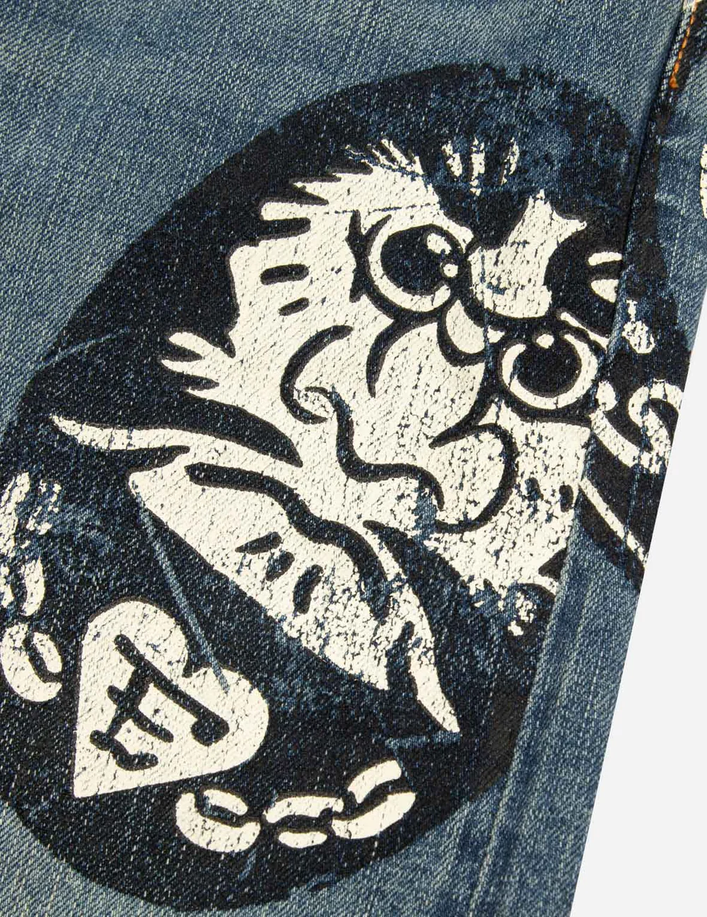 closeup shot of a pair of blue denim jeans with a distressed, vintagestyle graphic print. the print features a stylized cat or similar creature, a heart, and a chain, all done in a faded, whiteondark style.  the denim itself has a worn texture. the image focuses on the detail of the print and the texture of the fabric.
