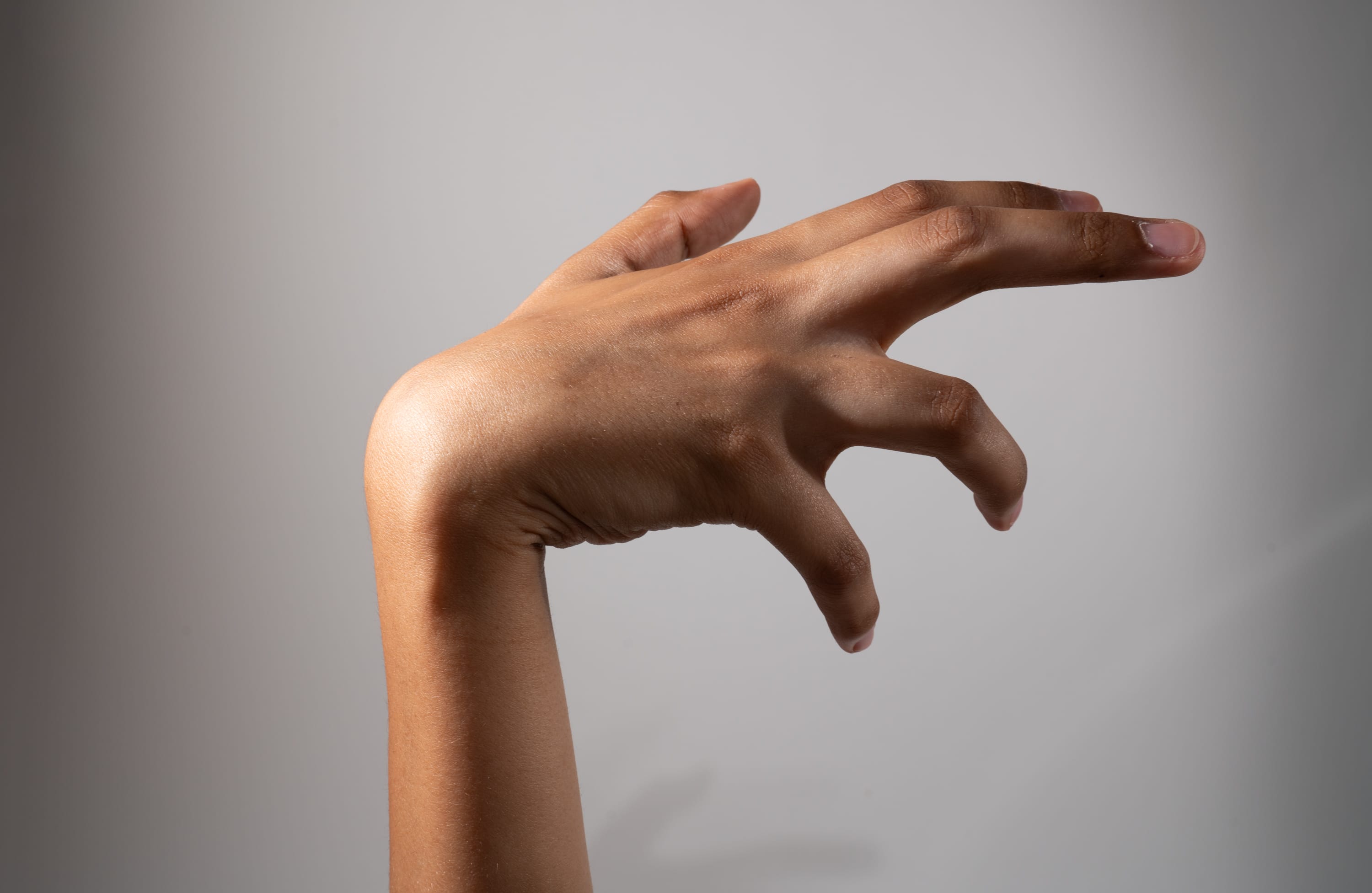 a closeup side view of a hand contorted into a clawlike pose against a light gray background. the hand, with its dark complexion, is illuminated by studio lighting, creating a dramatic effect.