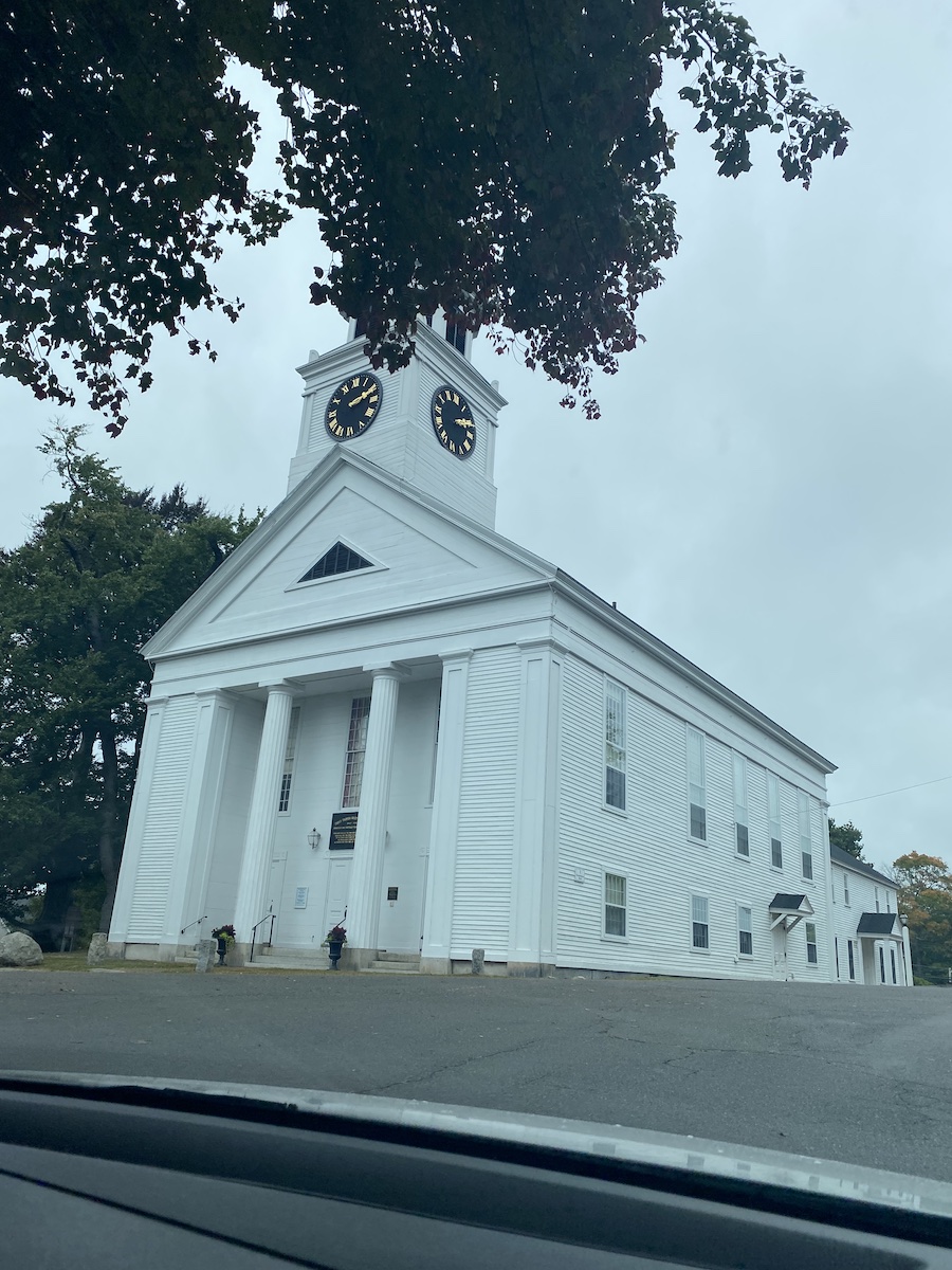 a lowangle shot of a large white church with a clock tower, columns, and several windows, with trees and overcast sky in the background.