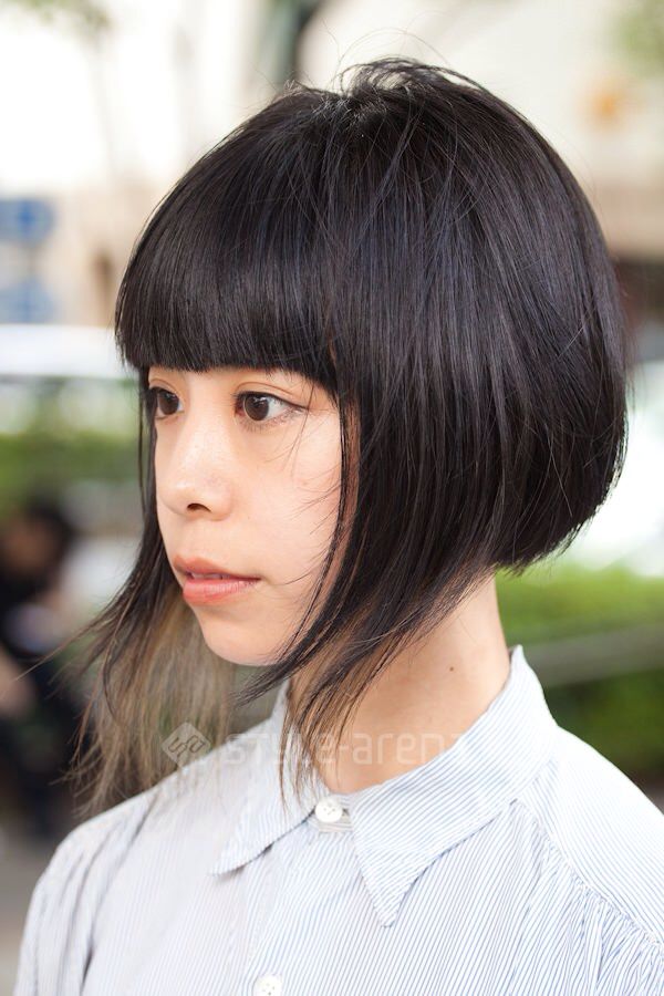 a close up shot of a young asian woman, with short black bob haircut and bangs, looking at the camera. she is wearing a white and blue striped collared shirt. the background is blurred and the image has a casual feel.