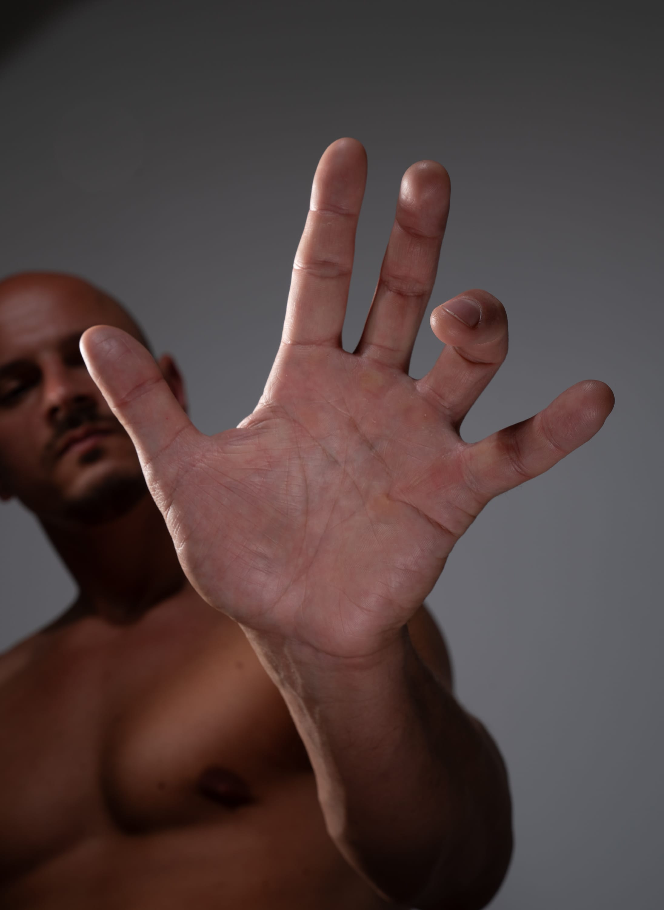 a closeup shot of a bald man with tan skin holding up his hand, palm out, in a gesture of halting or stopping. the focus is on the hand, with the man's face blurred in the background. the background is a neutral gray, and the lighting is soft and even.