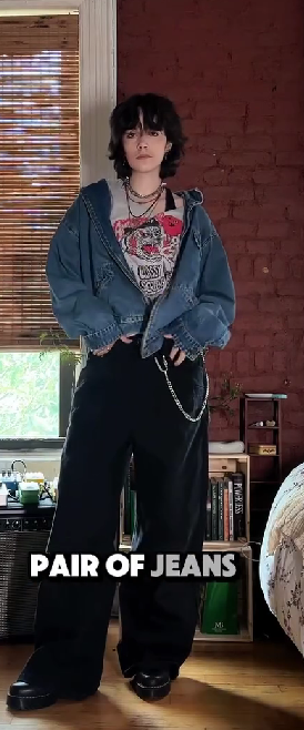 a young woman with short black hair is standing in a room wearing a blue denim jacket, a black and white graphic tshirt, black wideleg pants, a silver chain necklace, and black boots. she is leaning slightly forward with a relaxed look on her face.