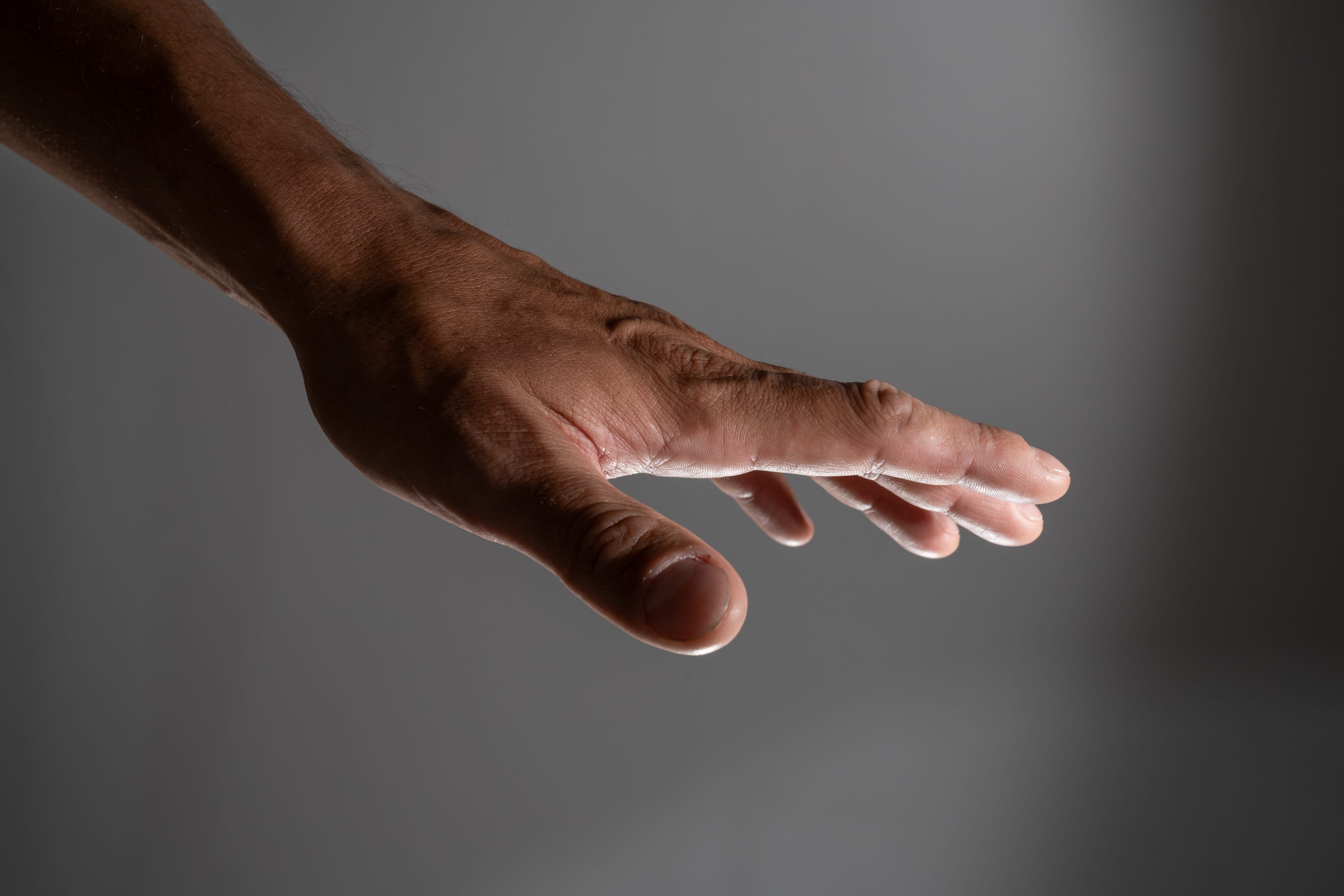 a closeup image of a man's outstretched hand against a gray background. the hand is reaching forward with fingers slightly spread, creating a sense of movement and anticipation. the lighting is soft and dramatic, highlighting the texture and details of the skin.