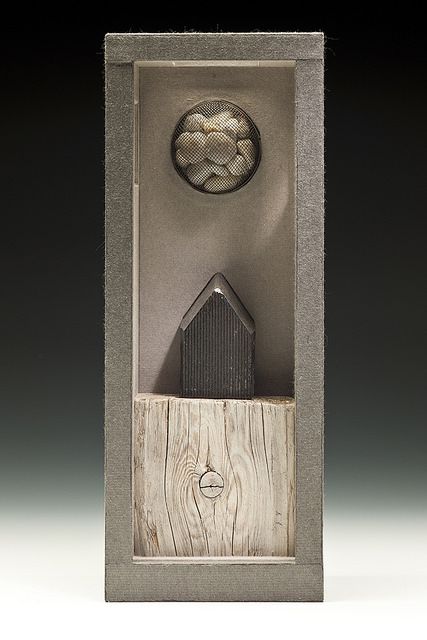 a vertical still life features a textured gray rectangular frame containing a mixed media assemblage. at the bottom, a section of weathered wood with a prominent knot serves as a base. above it, a small, dark, houseshaped object rests, likely made of a different material such as metal or a dark stone. in the upper portion of the frame, a circular wire mesh holds a collection of rounded, lightcolored stones. the overall color palette is muted, dominated by gray tones and natural textures. the artwork has a tactile quality, suggesting a blend of natural and manufactured elements.