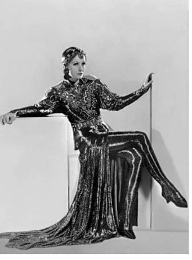 a woman dressed in a black, sequined gown poses sitting in a studio. she is wearing a long, elegant gown and her hair is styled in a chic bob with a jeweled headband. she is leaning forward with one leg extended, and the folds of her gown create a dramatic effect. the lighting is dramatic and creates a sense of depth and shadow. this image is a classic example of 1930s hollywood glamour.