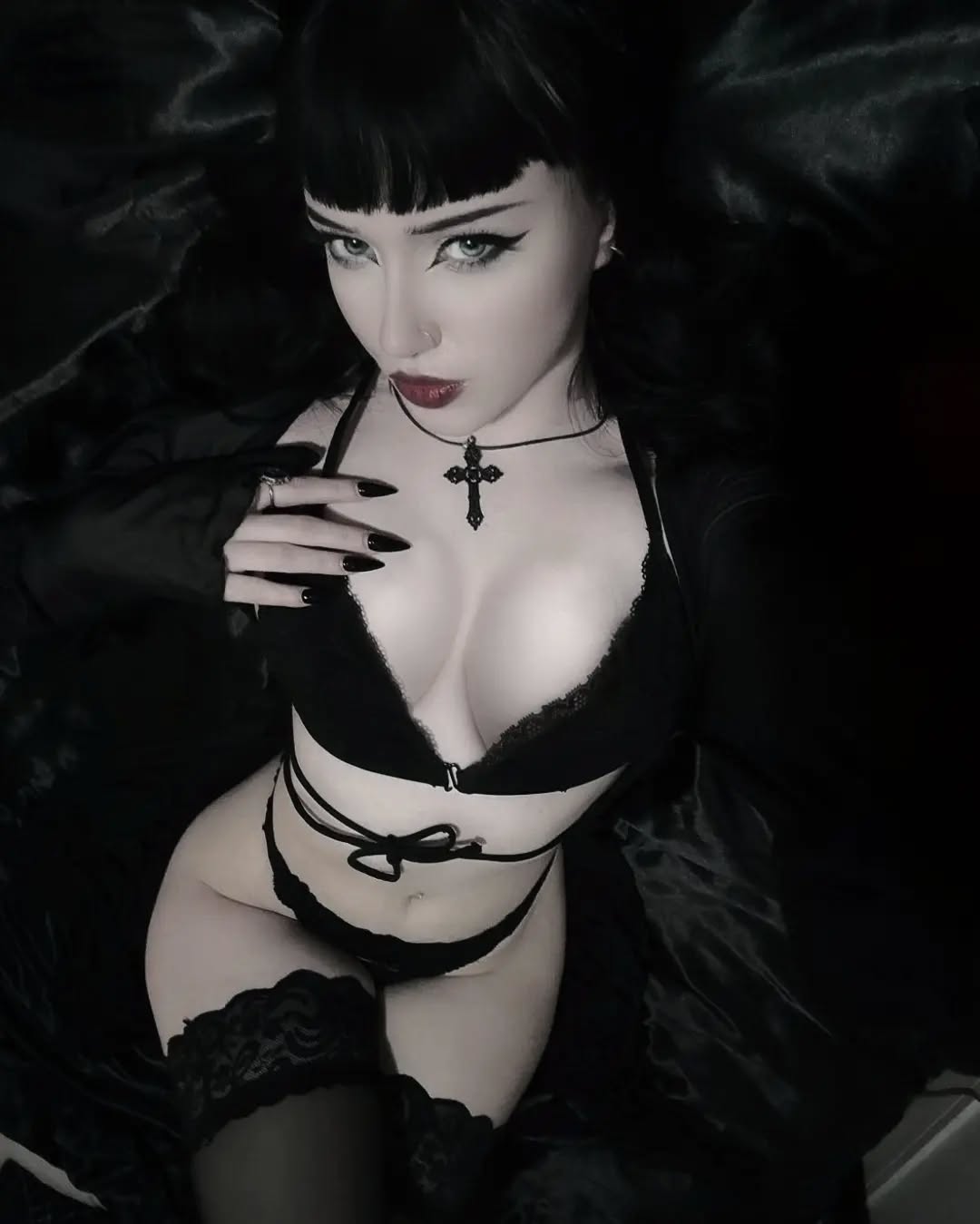 the image shows a young woman with long black hair styled with bangs. she's wearing dark makeup, including winged eyeliner and dark lipstick. her attire consists of black lingerie, a cross necklace, and possibly black stockings or thighhighs. the overall aesthetic is gothic and dark; the setting appears to be a dimly lit bedroom with black satin sheets. the photo is taken from a slightly elevated angle, offering a fairly closeup view of the woman's face and body. the lighting and color palette contribute to a moody and mysterious atmosphere. the woman's pose and expression are both seductive and somewhat melancholic.