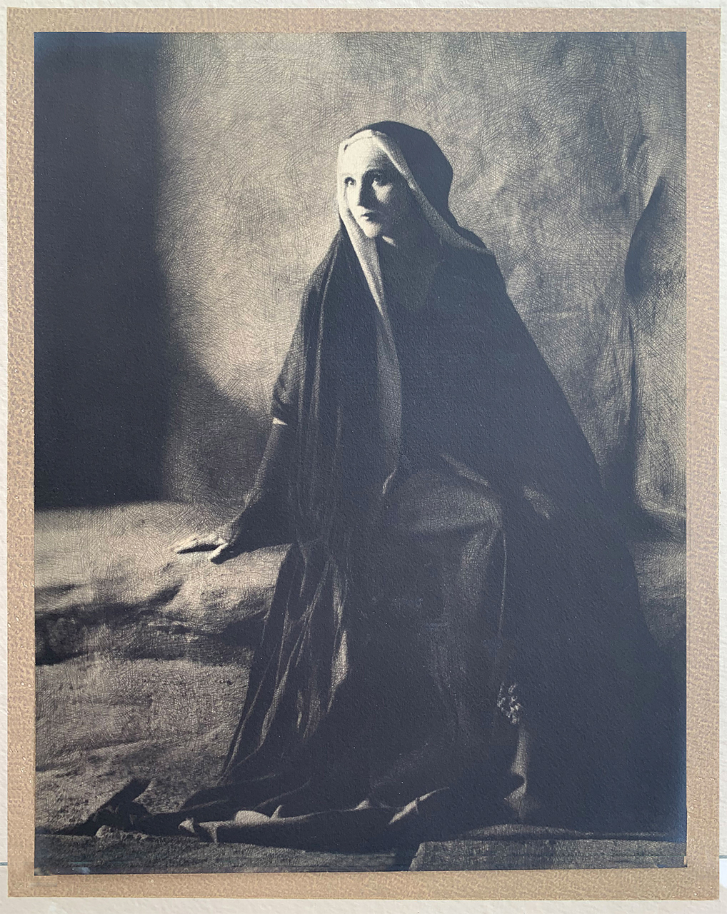the image is a striking black and white photograph, possibly a vintage print or reproduction of a vintage photograph, portraying a woman dressed in a dark, hooded robe reminiscent of medieval religious attire. she is seated on the ground, her pose contemplative and somewhat sorrowful. the lighting is dramatic, utilizing chiaroscuro—a strong contrast between light and shadow—to highlight her face and the folds of her clothing. the background is dark and textured, further emphasizing the figure in the foreground. the overall mood is somber and artistic, suggesting a possible religious or allegorical theme.  the photographic style and quality indicate a high level of skill and artistry.