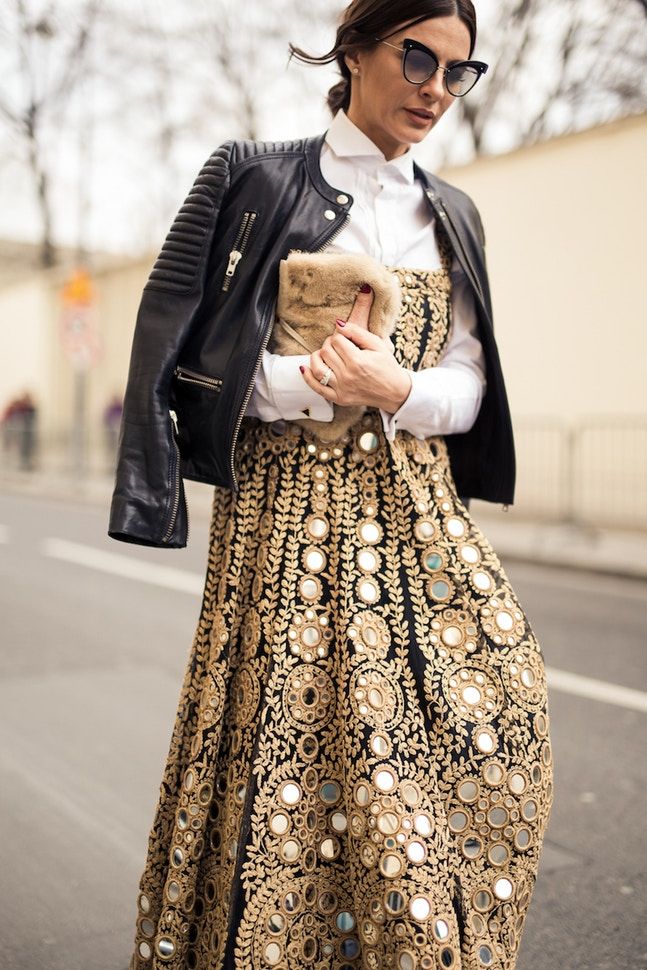 a stylish woman is captured in a street style photograph.  she's wearing a stunning black dress heavily embroidered with gold thread and small mirrors, creating a richly detailed and textured look.  over the dress, she wears a crisp white shirt and a black leather biker jacket with quilted shoulders. her accessories include sunglasses, a furry clutch, and rings. the setting appears to be a city street. the overall style is a sophisticated mix of bohemian and edgy elements.