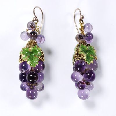 a closeup shot shows a pair of elegant dangle earrings. each earring features a cluster of amethystcolored beads, shaped and arranged to resemble a bunch of grapes.  these beads appear to be made of glass.  each grape cluster has a small gold setting with an enameled green leaf, adding a touch of natureinspired detail.  the earrings are delicately designed, suggesting a vintage or antique style. the overall color palette is rich with purples and greens against a neutral background.