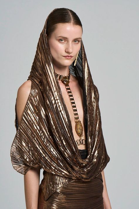 a woman in a gold metallic dress with a plunging neckline, and a gold chain necklace with a large stone pendant. she has her hair pulled back and is wearing minimal makeup. she is standing against a gray background.