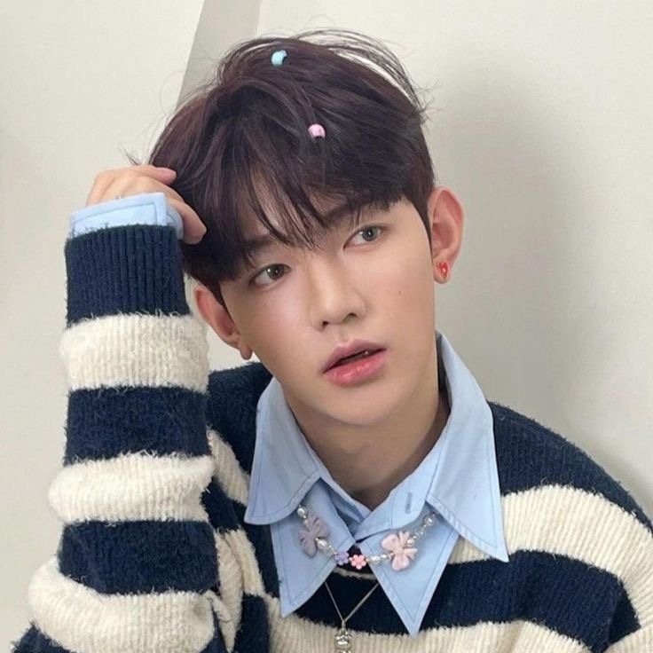 a young asian man is taking a selfie. he has short dark hair with colorful clips in it. he is wearing a blue and white striped sweater over a light blue collared shirt. he is also wearing a necklace with a flower pendant. the background is a plain white wall.