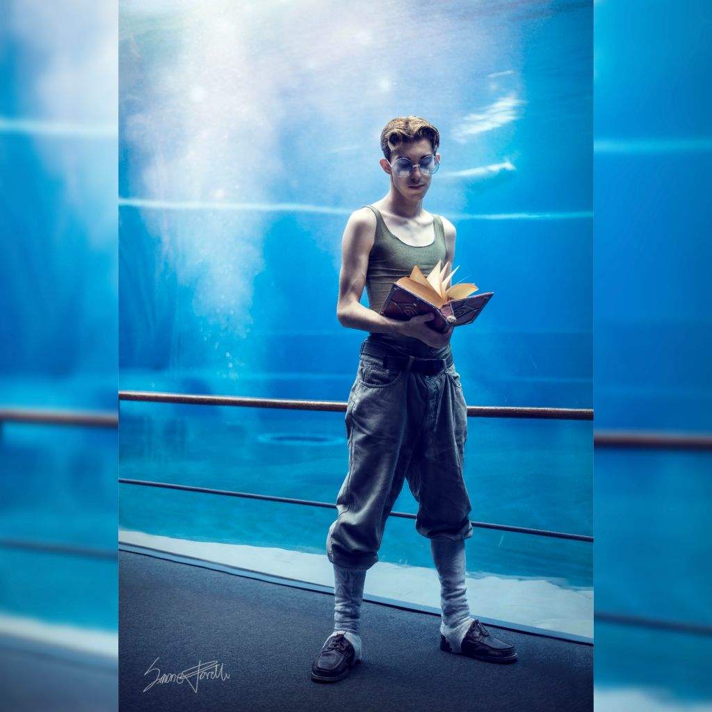 a man stands indoors next to a large aquarium, holding a book. he wears a tank top, trousers, glasses, socks, and shoes. the background is blue water with a cloudy effect.