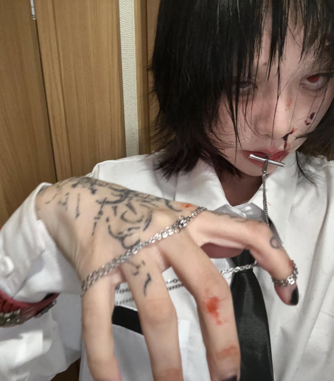 a person, presumably a woman, with short black hair, wearing a white shirt and a black tie. they have tattoos on their hand and are wearing rings and a chain. there appears to be blood or gore on their face and hand, and they have a metal cross in their mouth. the person is looking at the camera with a slightly creepy expression.