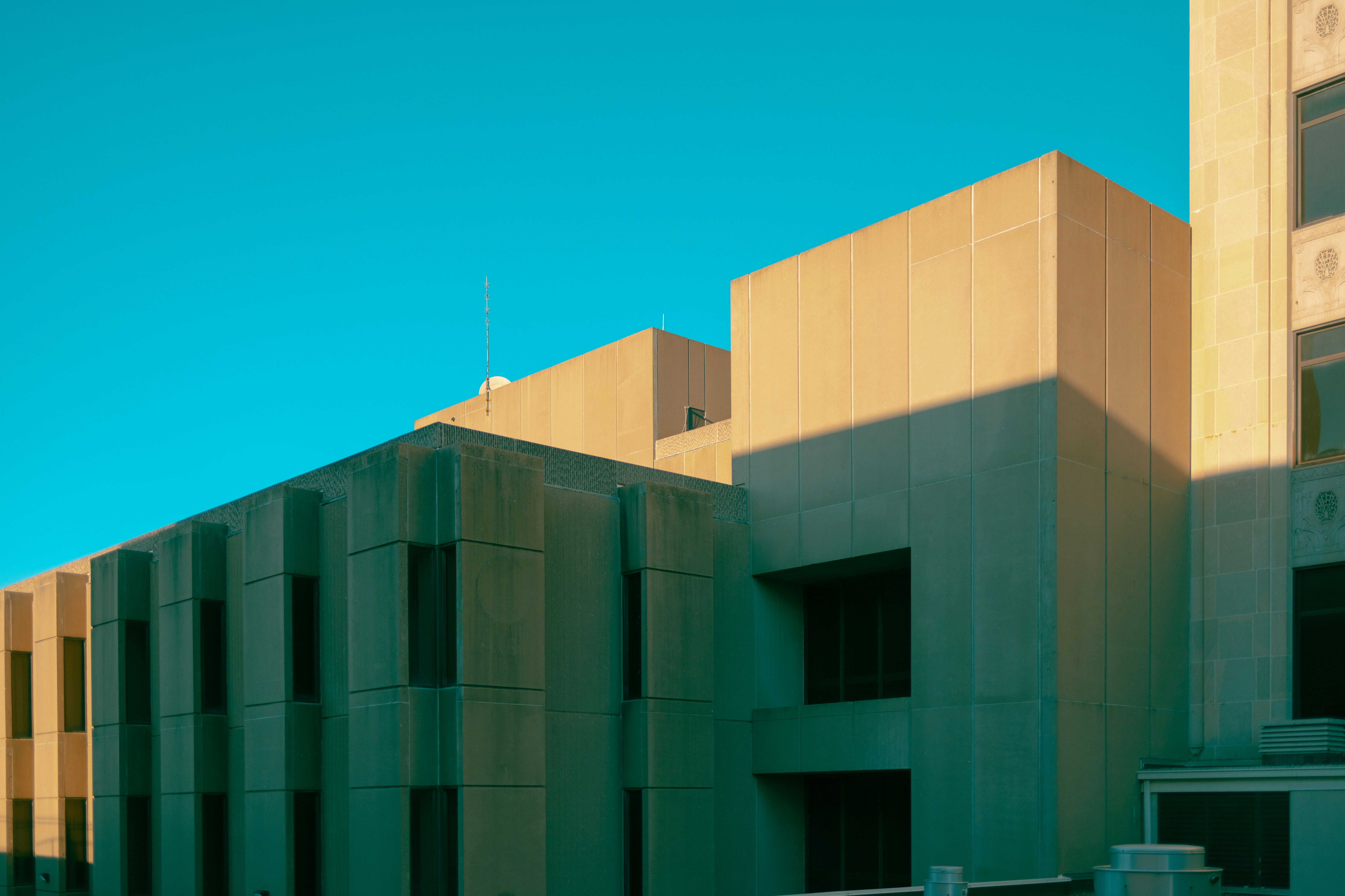 the image captures a portion of a modern building's facade against a vibrant blue sky. the building features a minimalist design with clean lines and geometric shapes, constructed primarily from concrete.  sunlight bathes the structure, casting long shadows that emphasize its threedimensionality and create a sense of depth.