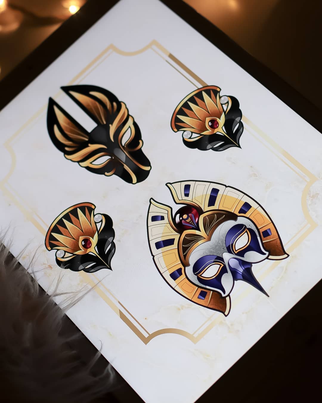 a digital drawing of four egyptian inspired mask designs displayed on a tablet. the design features two anubis masks and two pharaoh masks. the masks are designed with black, gold, and blue accents. the background is white with a gold border. the image is brightly lit with indoor lighting.