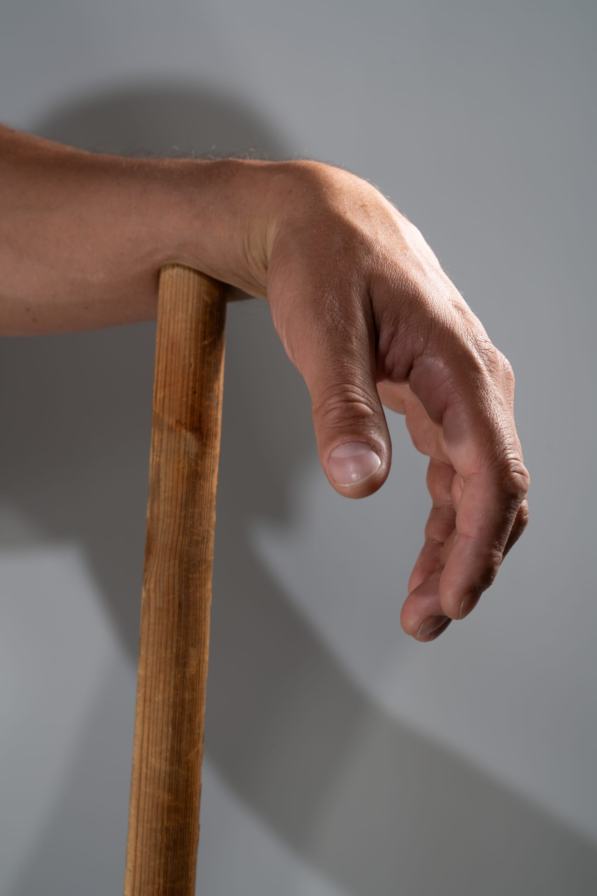 a closeup shot of a man's hand tightly gripping a wooden stick against a white background. the studio lighting creates distinct shadows, highlighting the texture of both the hand and the wood.