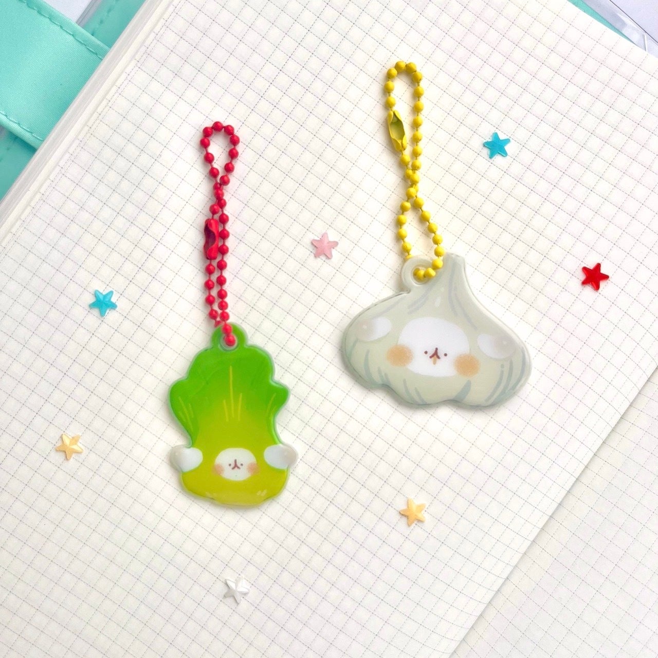 wo cute vegetable keychains are lying on a grid notebook. one keychain is shaped like a garlic clove and the other is shaped like a bok choy. both are pastel colored and have a cartoonish face on them. the keychains are attached to different colored chains (yellow and red). the notebook has some colorful stars scattered on it.