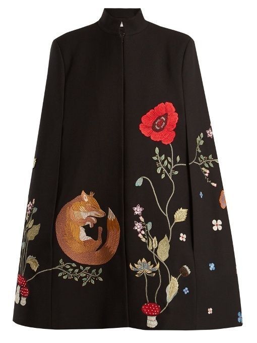 a black cape with intricate embroidery details. on one side, there's a curledup fox design, complete with foliage and mushrooms, while the other side features a large red poppy alongside other smaller floral patterns. the cape has a high collar and a structured silhouette.