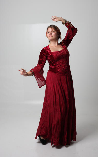 a fulllength shot of a woman in a flowing red velvet dress with long sleeves. she is standing on a white background, one arm raised above her head. her pose is elegant and graceful. the dress is detailed and appears to be of medieval or renaissance style. she is wearing jewelry such as bangles and earrings.  her expression is calm and slightly pensive, as she looks off to the side. the lighting and background are simple and clean, highlighting the dress and the woman's posture.