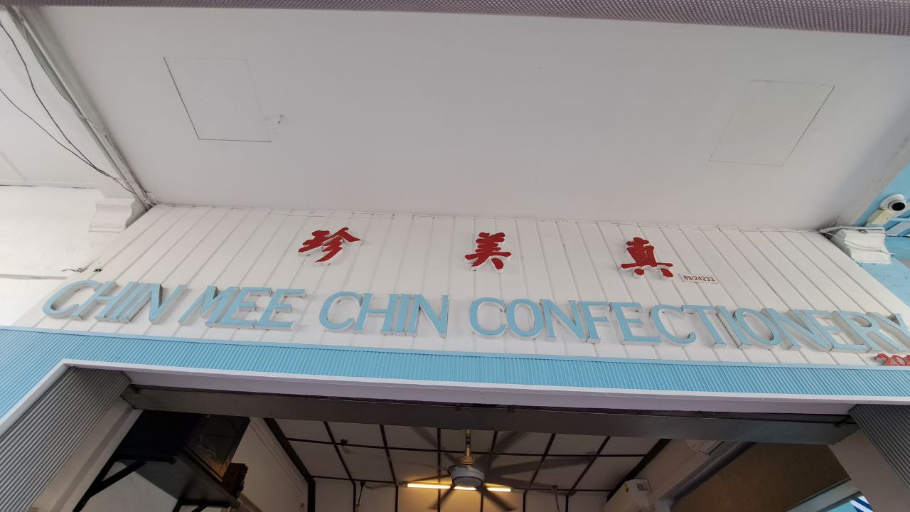 a storefront sign for a chinese bakery called chin mee chin confectionery. the sign is white with red chinese characters and blue letters. the shop is located in a building with a white facade and a blue awning.