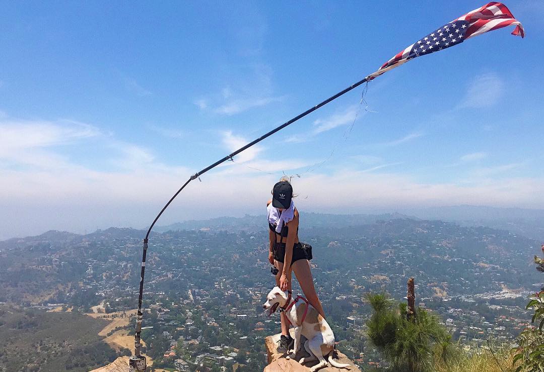a woman stands on a mountaintop with her dog, enjoying a panoramic view of a sprawling city below. an american flag is attached to a pole in the background, waving in the breeze. the sky is bright blue, with a few scattered clouds. the scene is serene and suggests a sense of freedom and adventure. the woman is wearing a black sports bra, black shorts, a black adidas hat, and sunglasses. her dog is a light brown and white pit bull mix.  the overall feel of the image is one of accomplishment and enjoyment of the outdoors.