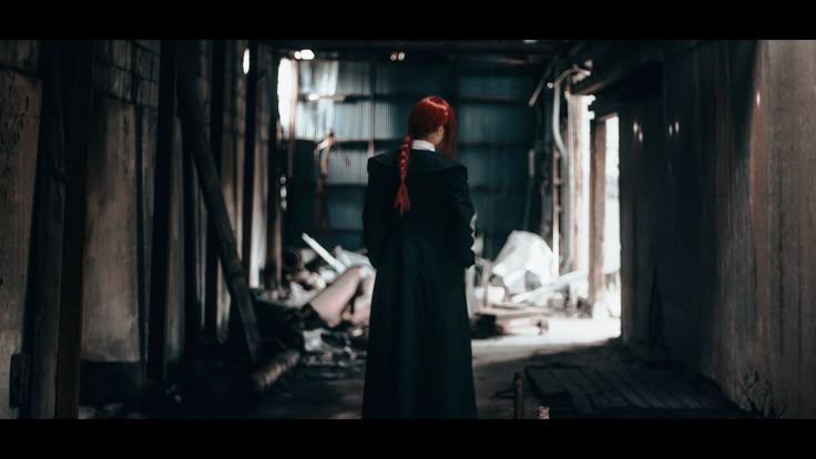 a person with red hair in a braid wearing a long black coat stands in a dark, ruined building filled with rubble. the person is facing away from the camera.