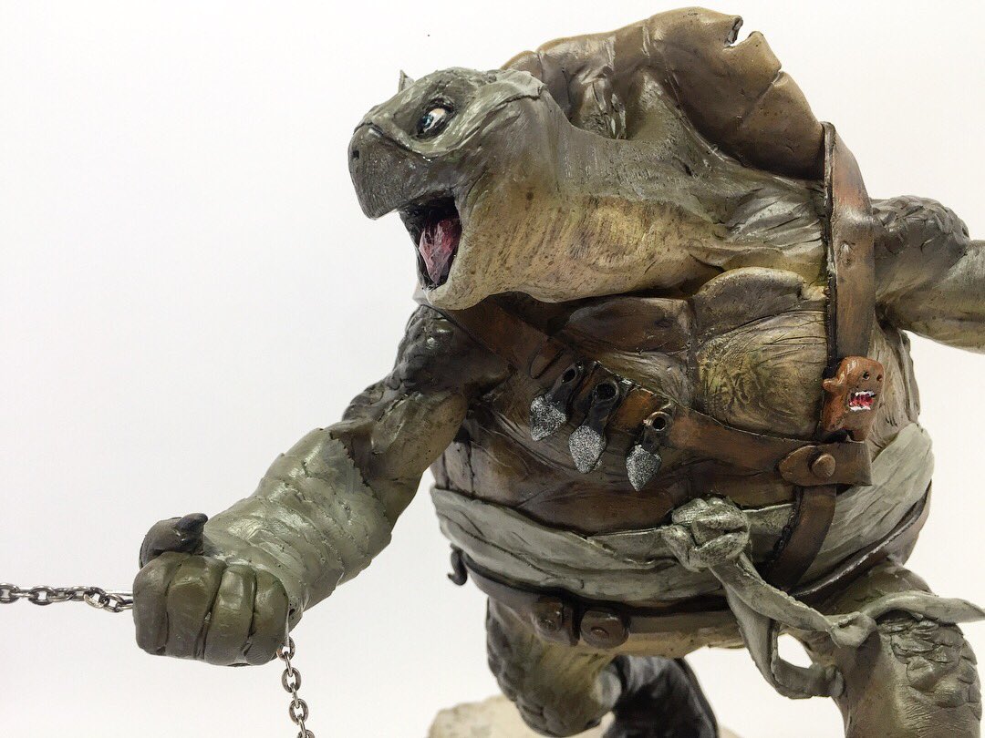 a figure of a humanoid turtle with an angry expression and open mouth. it has leather straps across its body, bandages on one arm, and holds a chain. there are weapons attached to the straps. the turtle has a shell on its back.