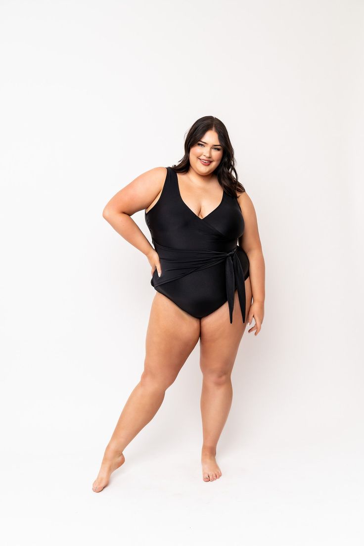 a plussize woman smiles confidently while wearing a black wrapstyle onepiece swimsuit against a white background.  the swimsuit is a dark solid color with a flattering vneck and a wrap detail at the waist. the woman is posing slightly sideways, showing her body in a relaxed and comfortable stance. the image promotes body positivity and confidence in swimwear.