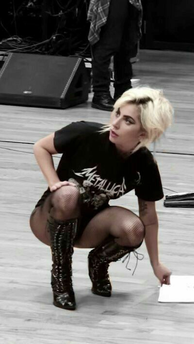 the image shows lady gaga backstage, possibly during a rehearsal or photoshoot. she's crouching on a grey floor, wearing a black metallica tshirt, fishnet stockings, and dark laceup boots.  the setting appears to be a stage area with audio equipment, cables, and speakers visible in the background. the image is in black and white, enhancing a slightly gritty and edgy aesthetic. a person in plaid shirt and dark pants is blurry in the background.  she appears to be looking down at something, possibly signing an autograph or working on something on the floor.