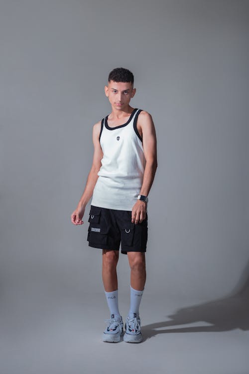a young man stands against a grey background wearing a white tank top, black cargo shorts, light blue sneakers, and white socks. he is posing for a fashion photograph.