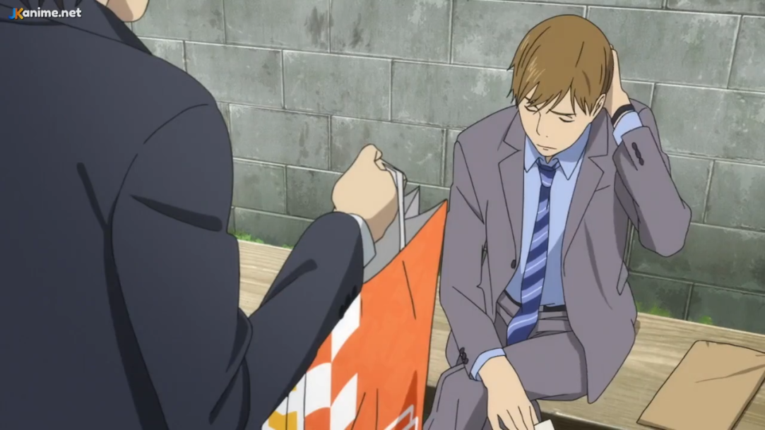 two men are having a serious conversation on a bench outside against a brick wall.  the man sitting is dressed in a gray suit with his head down, seemingly upset.  the other man is standing and holding a bag while facing away from the camera.  the scene is set during the daytime with natural light.