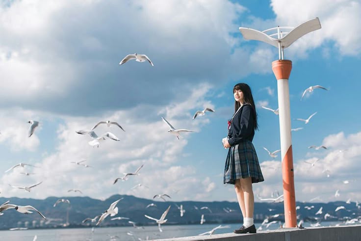 a young asian woman in a school uniform stands on an embankment near a body of water, surrounded by seagulls. the sky is blue with some clouds, and there is a street light nearby. in the background, there is a mountain range along the horizon.