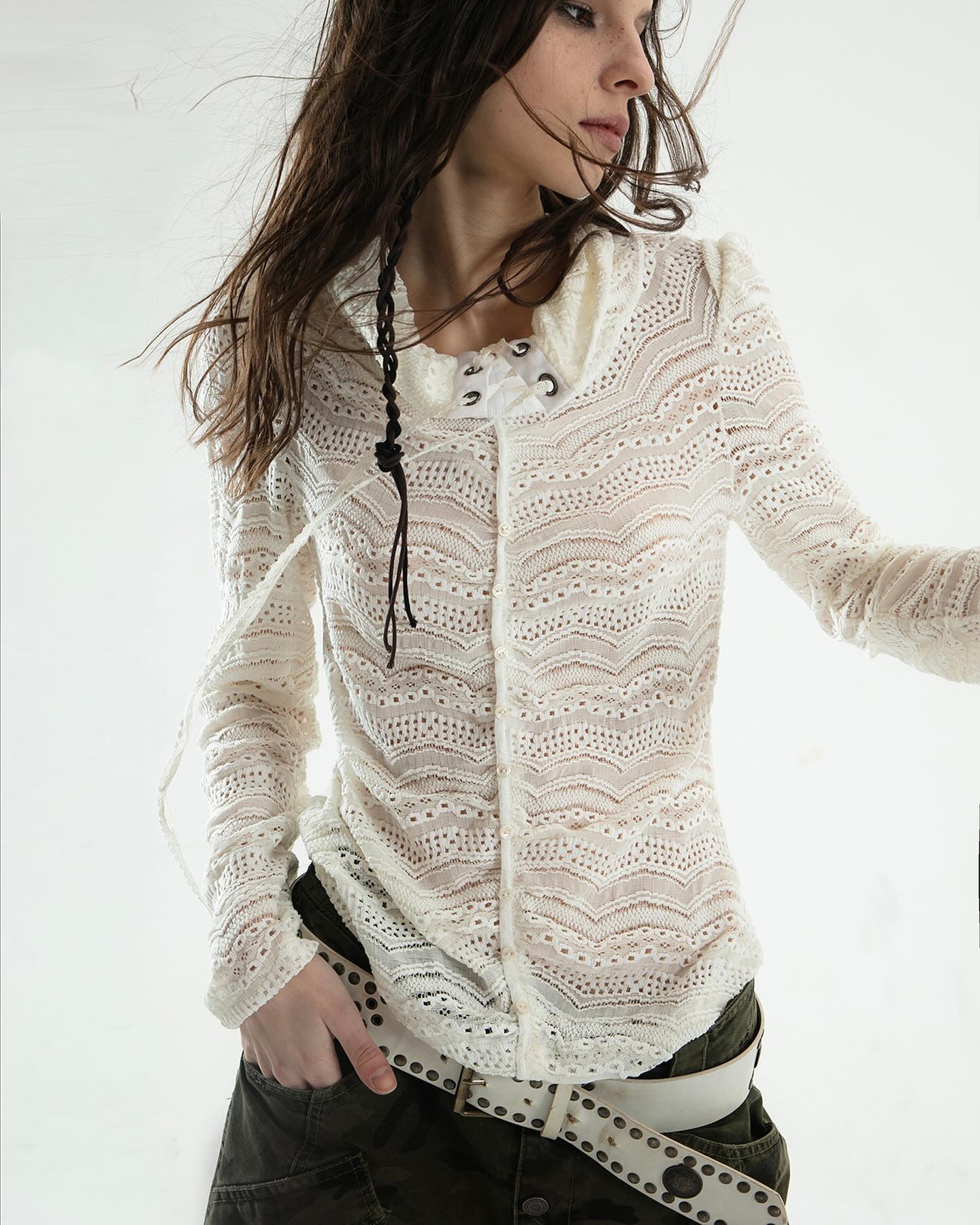 the image shows a young woman with long brown hair, styled in a braid, wearing a sheer, offwhite lace top with a high neckline and long sleeves. the top has a crocheted or knitted texture with lace detailing. the top is loosefitting. she is also wearing a green cargostyle pants and a white belt with studs. the background is plain white. the overall style is boho or vintageinspired. the photo is a fashion portrait, likely taken in a studio.