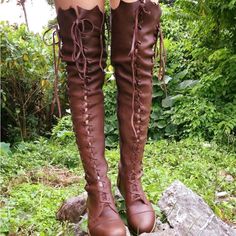 a woman is standing on a rocky surface, surrounded by greenery, showcasing a pair of brown laceup overtheknee leather boots. the boots have a vintage and bohemian aesthetic. the lighting is natural, highlighting the texture of the leather and the natural surroundings.
