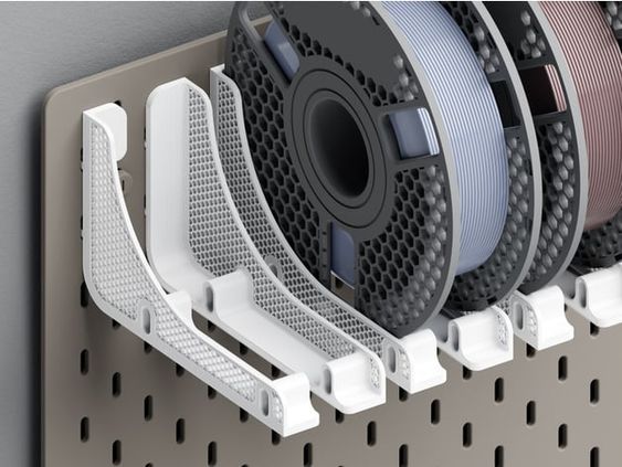 a closeup image of a wallmounted filament holder with three spools of filament for a 3d printer. the filament holder is made of white plastic and has a grid pattern. the filament spools are black and have a hexagonal pattern. the spools are mounted on the wall holder using pegs that fit into the holes in the holder. the holder is mounted on a grey pegboard wall.