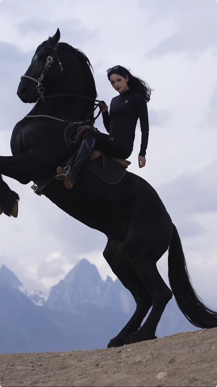 a woman rides a black horse that is rearing up on its hind legs, set against a backdrop of mountains and a cloudy sky. the woman is wearing a dark, longsleeved outfit and the horse has riding gear. the scene evokes a sense of power and the outdoors.
