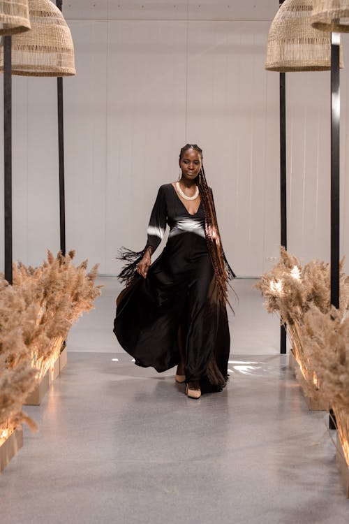 a black woman model with long braids walks down a runway wearing a long black dress with white accents and fringe. the dress is elegant and stylish, and the model's pose is confident. the runway is set in a minimalist, modern interior with pampas grass decor and unique lighting fixtures. the overall atmosphere is sophisticated and chic, reflecting high fashion.