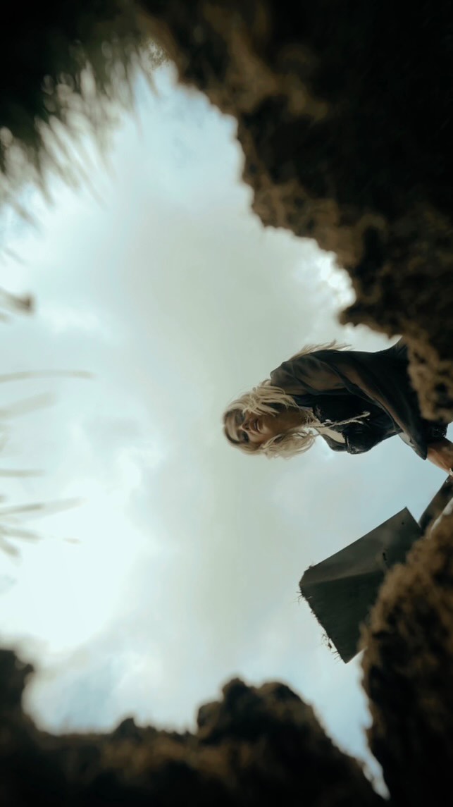 a lowangle shot from inside a hole or grave, looking up at a woman holding a shovel. the sky and clouds are visible in the background. the woman is wearing dark clothing and has blonde hair. the perspective gives a sense of being trapped or buried.