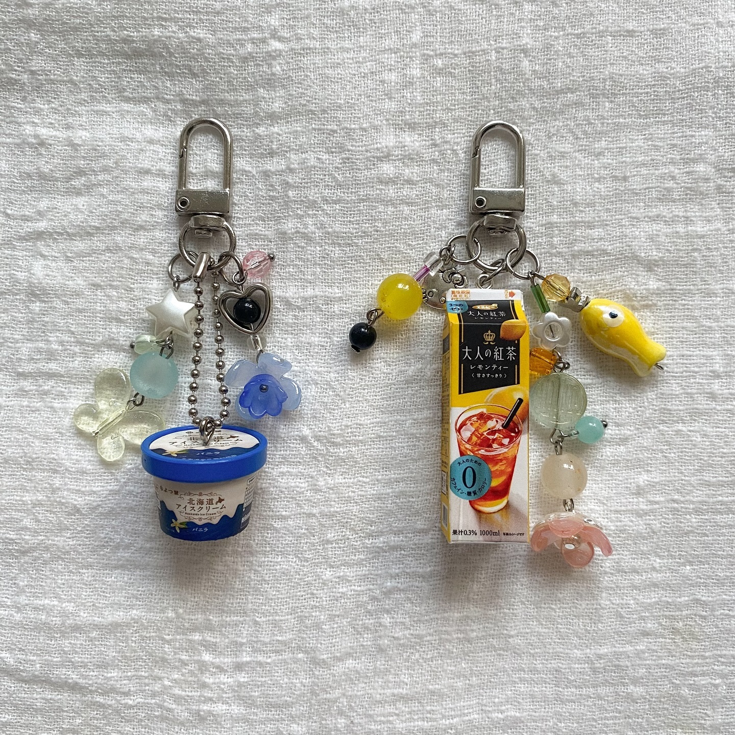 two keychains with miniature charms on a white textured surface. the keychain on the left has an ice cream container charm, flower charms, a heart charm, star charm and colored beads. the keychain on the right has a juicebox charm, a yellow fish charm, flower charm, and colored beads.