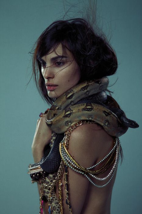 a closeup portrait of a woman with a captivating expression, draped in a snake, her body adorned with intricate jewelry. the soft lighting and moody atmosphere create a sense of elegance and mystique. the woman's gaze is directed downward, conveying a hint of mystery and allure.