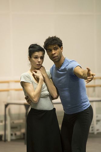 a woman and a man are dancing in a studio. the woman is wearing a gray leotard and a black skirt. the man is wearing a blue shirt and black pants. the woman is looking scared.