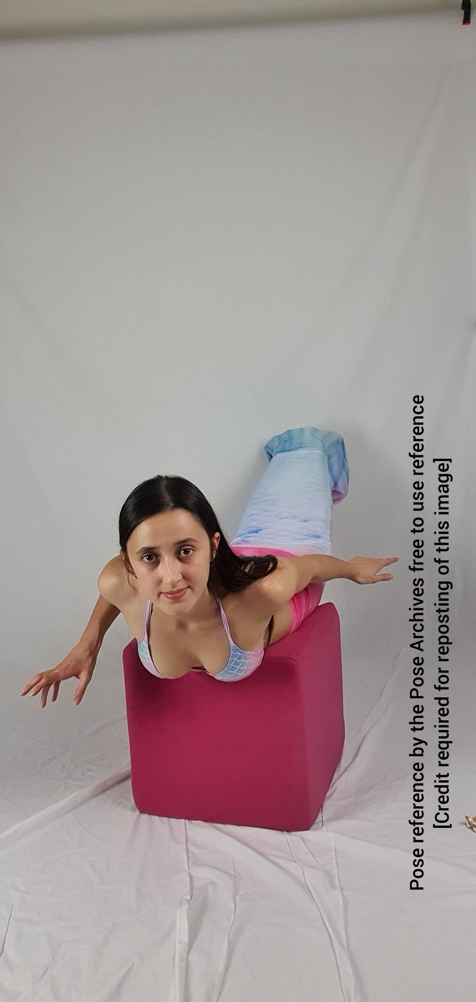 a young woman with long dark hair is positioned on her stomach on a pink cube. she's wearing a bikini top and a mermaid tail, her arms outstretched in a pose reminiscent of swimming. the backdrop is a plain white studio setup.
