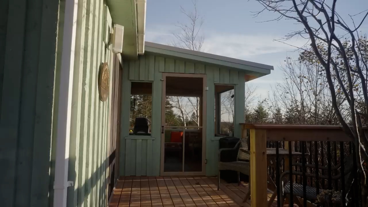 a wooden deck with railing leads to a screenedin porch attached to a light green house. the porch has a door and windows, offering a view of the surrounding trees. there is patio furniture on the porch, including a chair. the trees have mostly bare branches, suggesting it is autumn. the sky is blue, and the sun is shining, creating a warm and inviting atmosphere.
